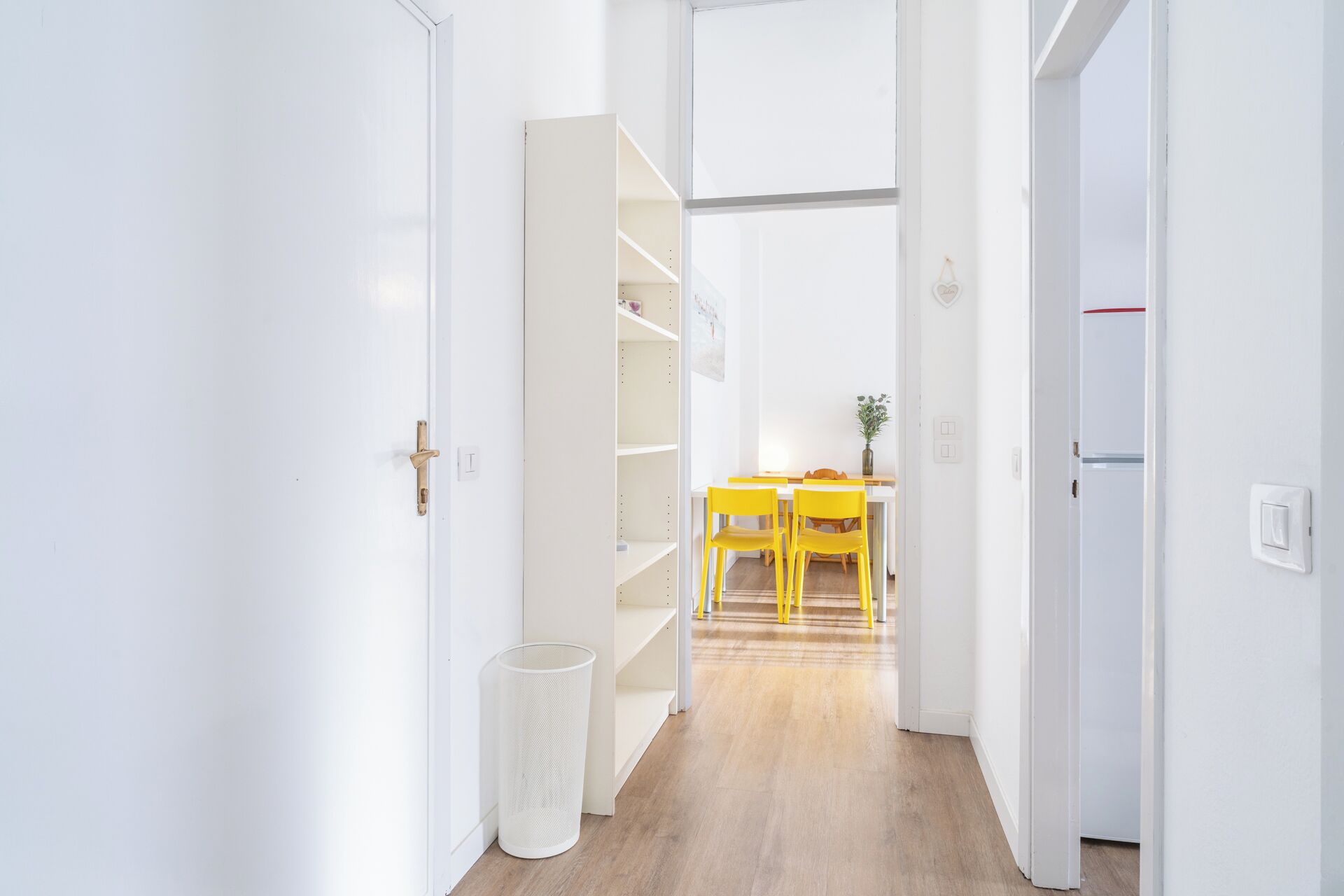 Un couloir aux murs blancs, doté d’un plancher en bois et comprenant un coin repas avec des chaises jaunes et une table.