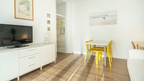A modern living room with a flat-screen TV, white furniture, a dining area with yellow chairs, and a beach scene painting.
