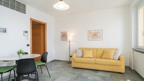 Una sala de estar moderna con un sofá amarillo, una mesa de comedor con sillas, una lámpara de pie y un ventanal grande.