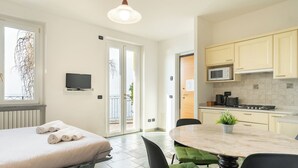 Una habitación de hotel moderna con cama, televisor, zona de comedor con mesa y sillas, y cocina equipada con armarios y electrodomésticos.