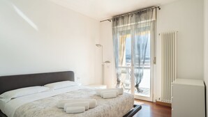 A bedroom with a bed, a nightstand, a lamp, a radiator, and a view of the outside through a window with sheer curtains.