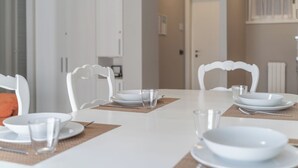 Ruokapöytä on katettu valkoisilla lautasilla, kulhoilla ja laseilla, ja sen ympärillä on valkoisia tuoleja, joiden selkänojissa on koristeellisia yksityiskohtia.