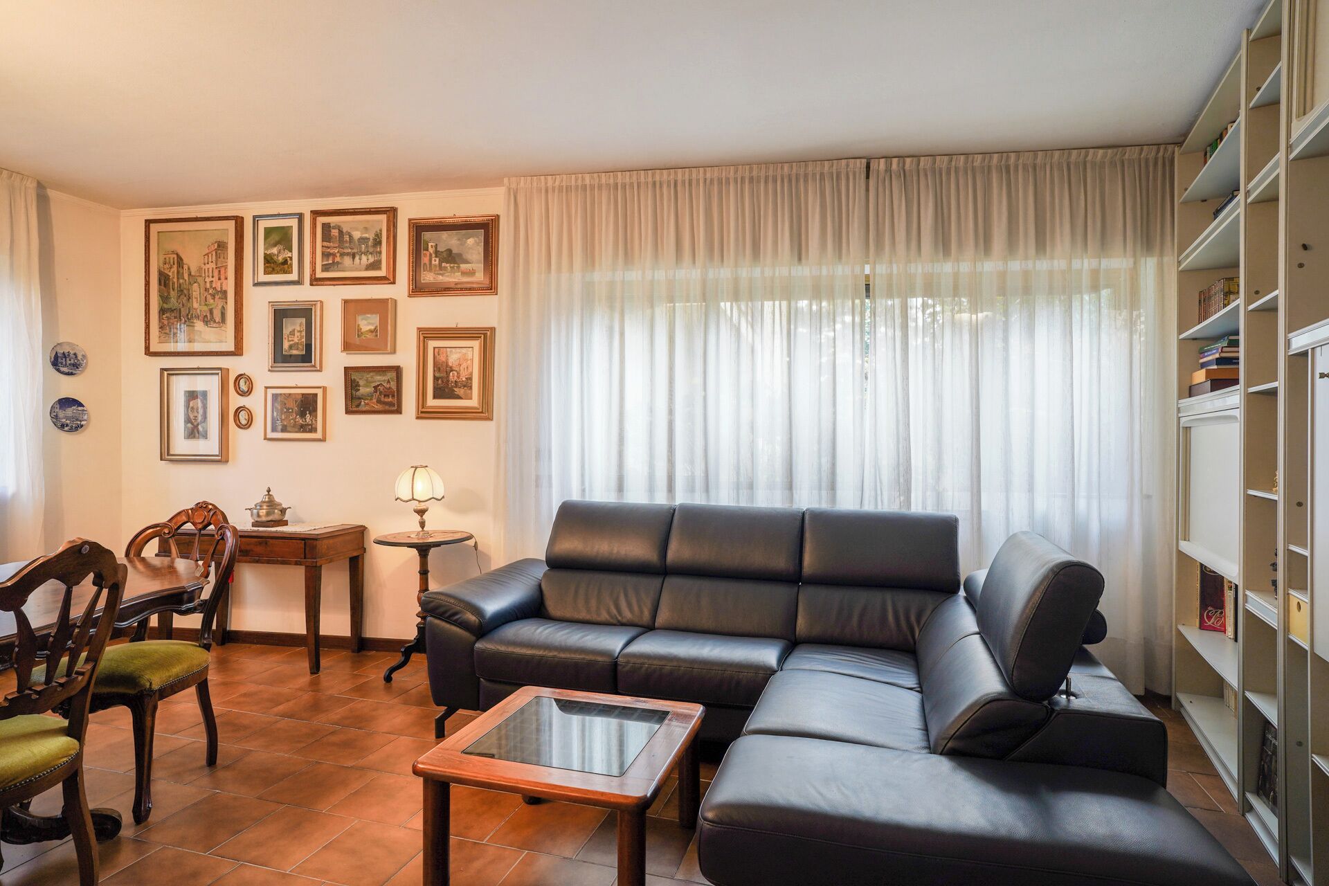 Een woonkamer met een zwarte leren bank, een houten salontafel en een eethoek met stoelen. Op de muur hangen ingelijste foto’s en een boekenkast zit vol boeken.