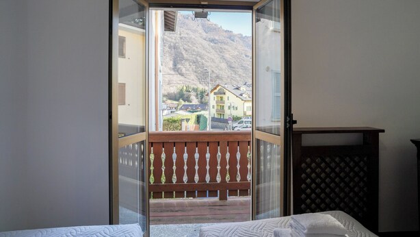 Een kamer met een bed, een houten balkon met een leuning en uitzicht op een berg en gebouwen.