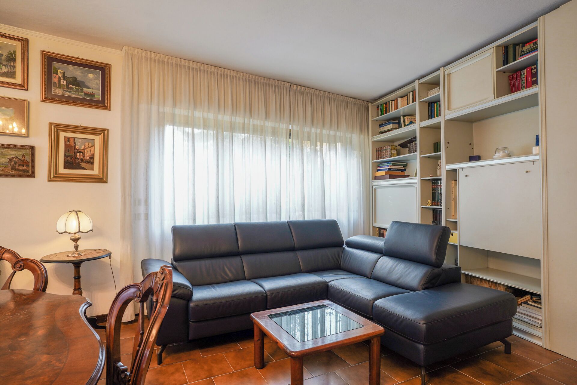Ein Wohnzimmer mit einem schwarzen Ledersofa, einem Couchtisch aus Glas, einem Holzesstisch mit Stühlen, einer Lampe und einem Bücherregal voller Bücher.