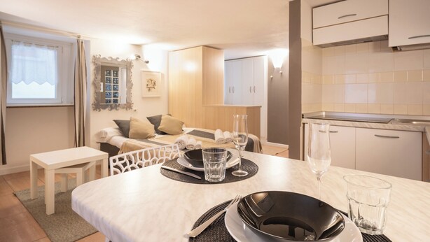 A modern, well-lit room with a dining table set for two, a sofa with cushions, a small table with a lamp, and a kitchen area with cabinets and a countertop. - Bellano Studio 2 - Studio Apartment, Sleeps 2 (Bellano)