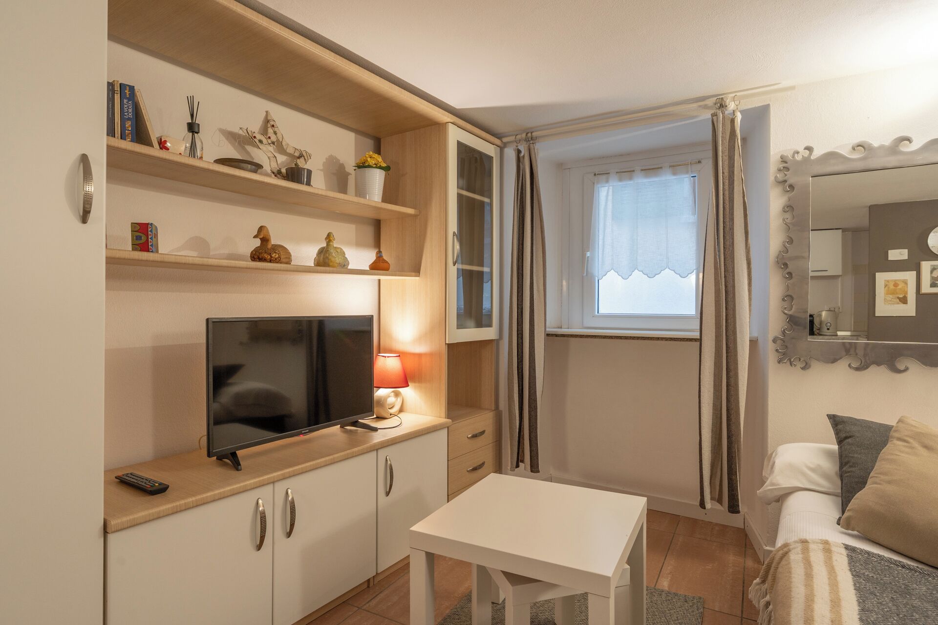 Un espace de vie compact comprenant un téléviseur, une petite table et un canapé. On y trouve des étagères ornées d’objets décoratifs et une fenêtre avec des rideaux.