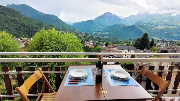 A balcony with a wooden table set for two, overlooking a mountainous landscape with houses and greenery.