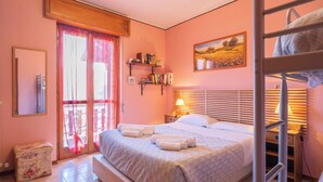 A bedroom with a bed, a bedside table with a lamp, a shelf with books, a window with curtains, and a bunk bed in the corner.