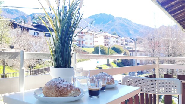 Un balcón con una mesa dispuesta para el desayuno, que incluye un pastel, café y una planta en maceta.