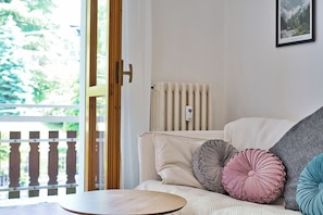 A room with a sofa, a round table, a radiator, and a window with a view of greenery.