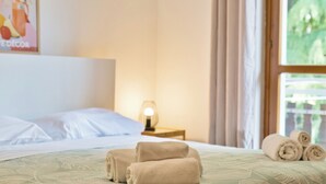 Una cama bien tendida con toallas enrolladas, una lámpara de noche y una ventana con cortinas.