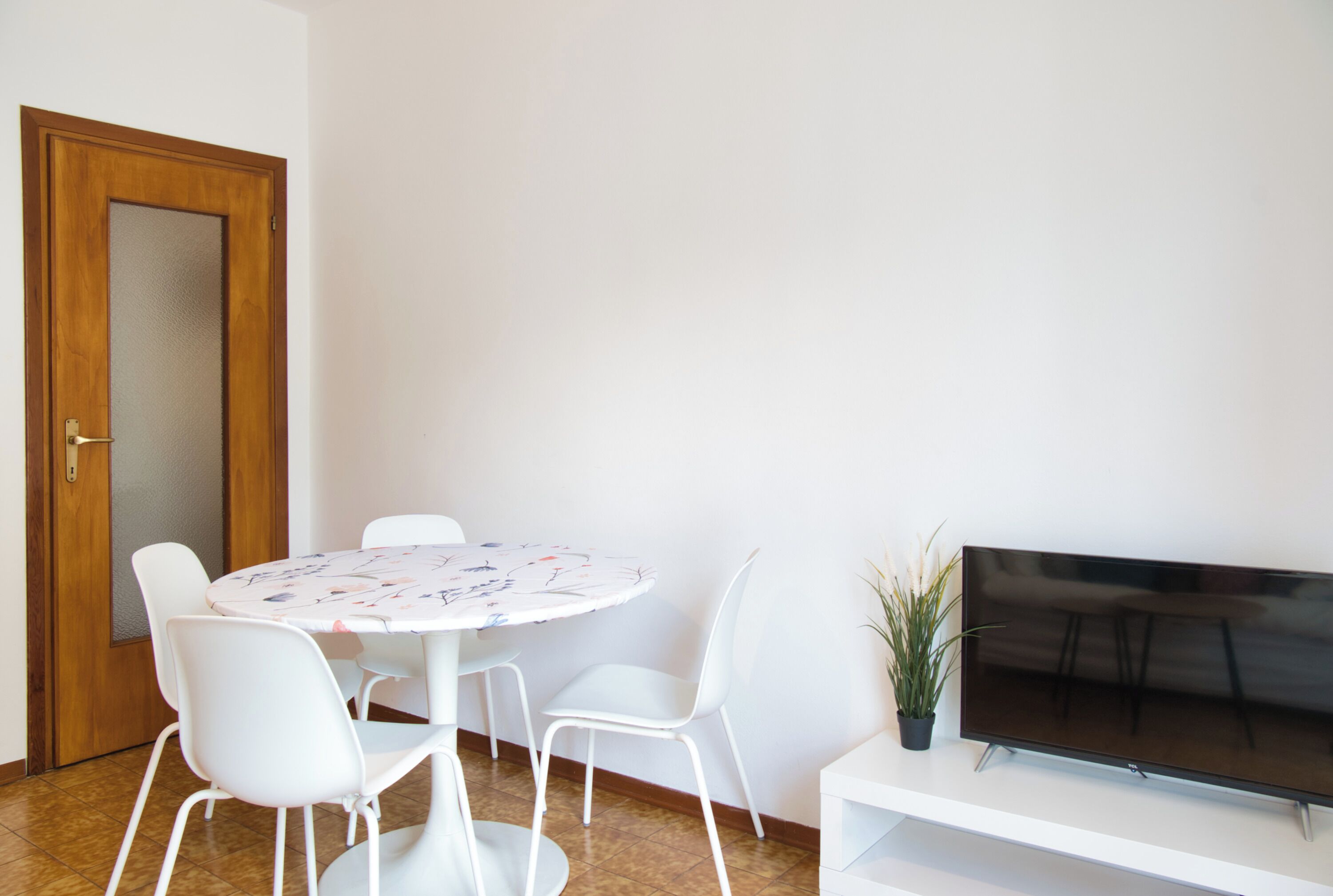 A room with a round table, white chairs, a wooden door, a flat-screen TV on a stand, and a potted plant.