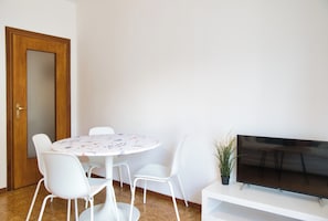 A room with a round table, white chairs, a wooden door, a flat-screen TV on a stand, and a potted plant.
