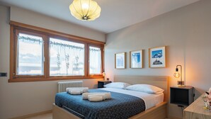Ein Schlafzimmer mit Bett, Heizkörper, einem Fenster mit Vorhängen, einer Wandleuchte und gerahmten Bildern.