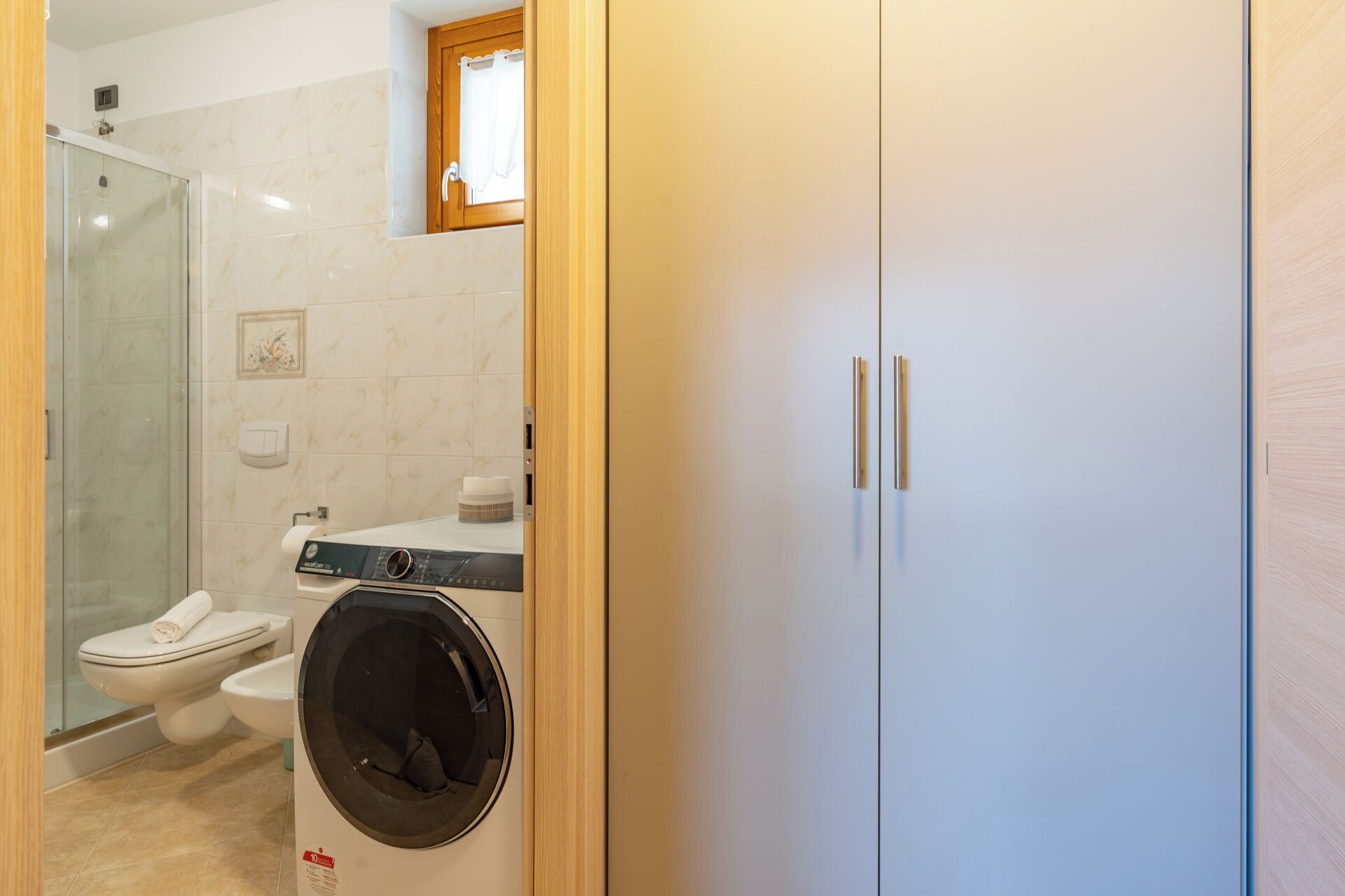 A bathroom with a glass shower enclosure, a toilet, a washing machine, and a wooden wardrobe.