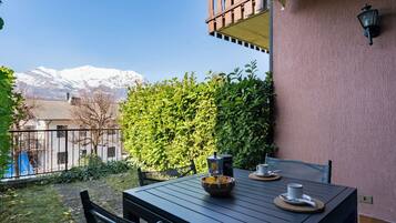 Une table de patio avec une machine à café, des tasses et un bol, le tout placé devant un paysage de montagne et d’une haie.
