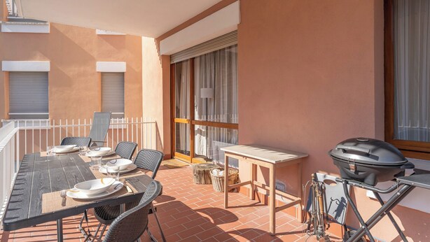 Un patio al aire libre con una mesa de comedor dispuesta para cuatro personas, rodeada de sillas de mimbre. Una parrilla se encuentra a la derecha.