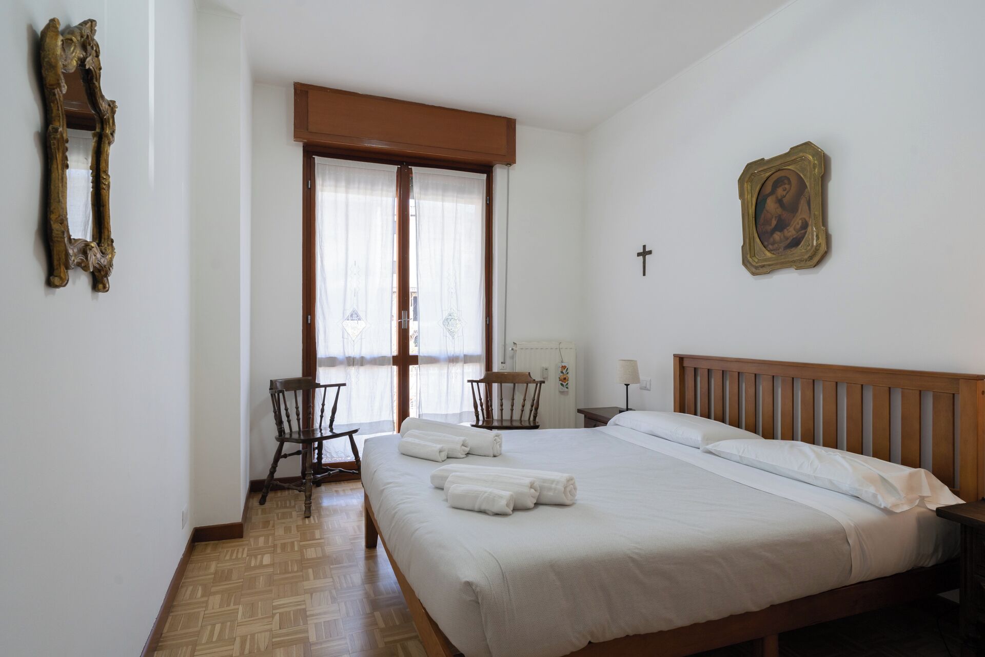 Un dormitorio con una cama grande, dos sillas, un cabecero de madera, una ventana con cortinas, un radiador y una cruz colgada en la pared.