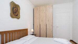 Un dormitorio con cabecera de madera, cama con ropa blanca, mesita de noche con lámpara, armario y una puerta.
