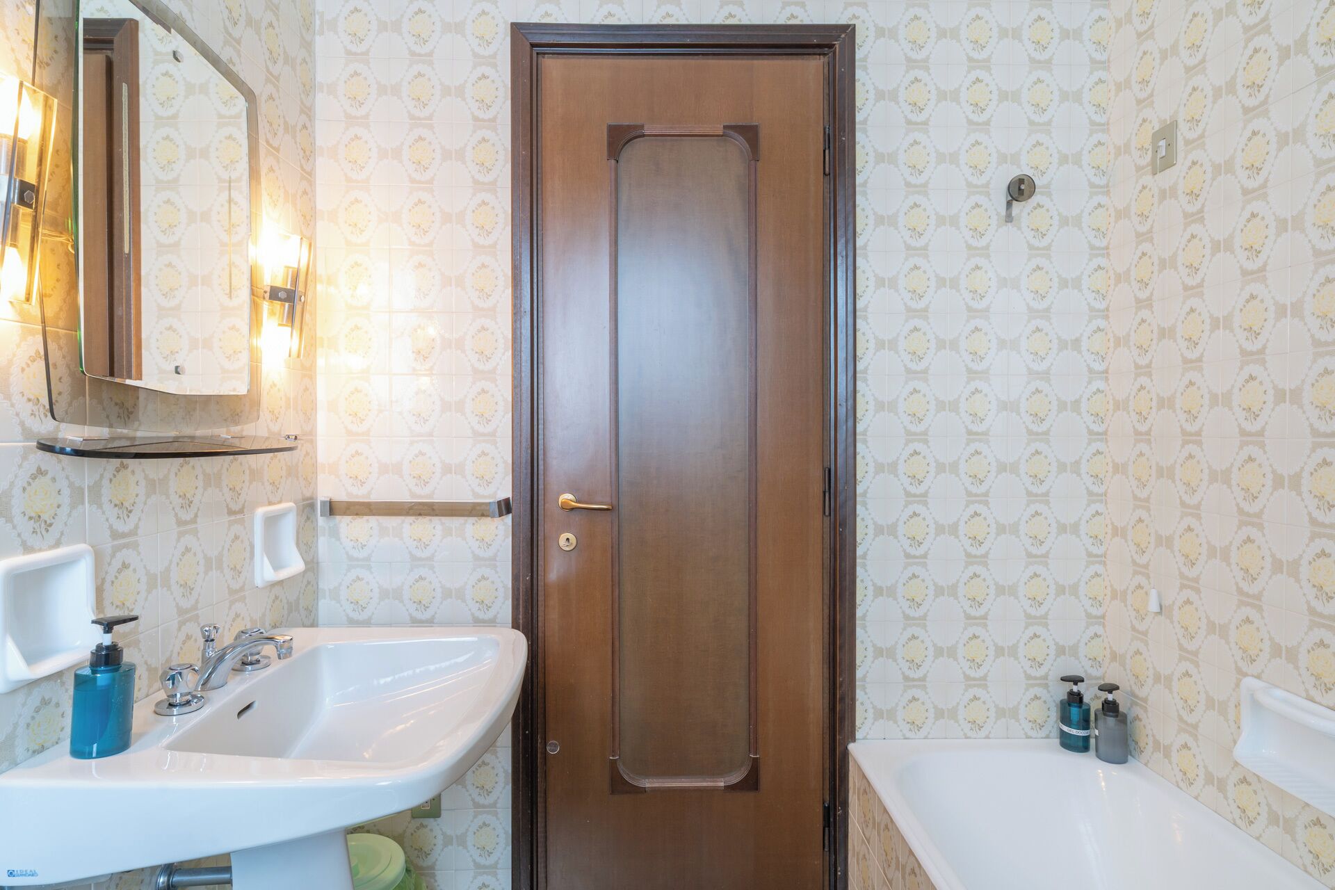 Une salle de bain avec une porte en bois, deux lavabos, un miroir et un papier peint à motifs.