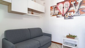 Een grijze bank in een kamer met een witte muur, een witte plank met een mand, en een kunstwerk met de tekst 'home sweet home'.