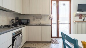 Een moderne keuken met een gasfornuis, magnetron en oven. Er is een spoelbak met een kraan, een raam met gordijn en een deur met een welkomstbord.