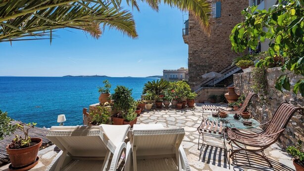 A terrace with lounge chairs and a table overlooking the sea, surrounded by potted plants and a stone building. - Seaside Neoclassical Property in Vaporia Ermoupoli (Syros)