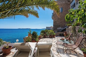 A terrace with lounge chairs and a table overlooking the sea, surrounded by potted plants and a stone building. - Seaside Neoclassical Property in Vaporia Ermoupoli (Syros)