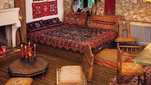 A rustic room with a stone wall, wooden beams, a large bed with patterned bedding, a wooden chair, and a small table with candles. - 260m2 Unique Mountain House in Lilea Parnasos (Delphi)