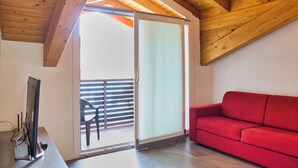 A room with a wooden ceiling, a sliding glass door leading to an outdoor area, a red sofa, and a desk with a computer and a phone. - Residence San Rocco - Appartmento Resegone (Cremeno)