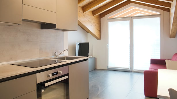 A modern kitchen with a built-in oven and a sink, wooden ceiling beams, and a sliding glass door leading to a bright room with a red couch. - Residence San Rocco - Appartmento Resegone (Cremeno)