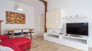 Ein modernes Wohnzimmer mit Flachbildfernseher, einem Esstisch mit Stühlen, einem roten Sofa, einem farbenfrohen abstrakten Gemälde und einer weißen Unterhaltungseinheit mit Regalen.