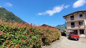 Ein zweigeschossiges Gebäude mit einem Balkon, ein rotes Auto davor geparkt und ein Berg im Hintergrund.