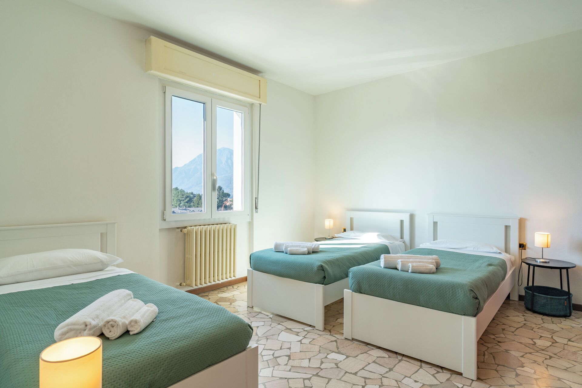 Et hotelværelse med to senge, et vindue med udsigt til et bjerglandskab, en radiator og et lille bord med en lampe.