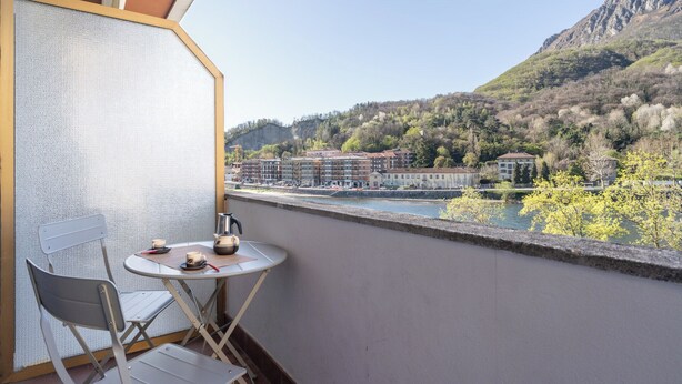 Ein Balkon mit Tisch und Stuhl, der auf einen Fluss und Gebäude in einer Berglandschaft blickt.