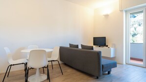 Ein modernes Wohnzimmer mit grauem Sofa, weißen Sesseln, einem Fernseher auf einem weißen Schrank und einem Balkon mit Blick.