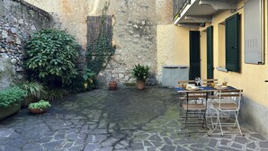 Un patio con suelo de piedra, plantas en macetas, una mesa con sillas y una ventana con contraventanas.