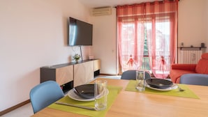 A dining area with a table set for two, a television mounted on the wall, and a balcony with a view.