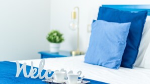 Un lit bien fait, avec une literie bleue, une table de chevet avec une lampe, une petite plante en pot et un panneau d’accueil.