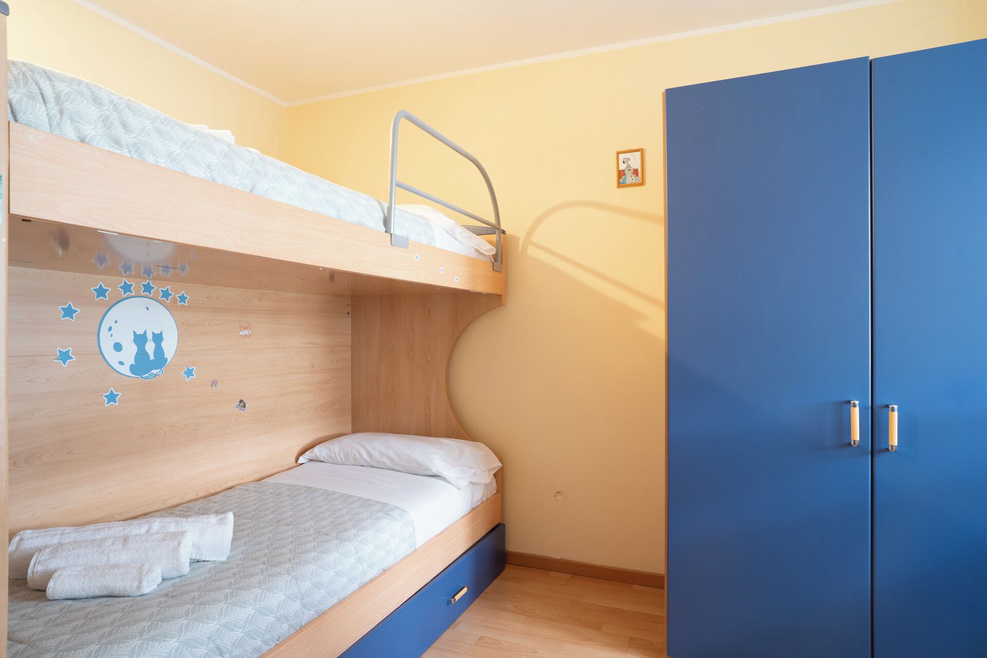 Un lit superposé avec un motif de ciel étoilé sur le mur, un lit simple avec un oreiller et une armoire bleue.