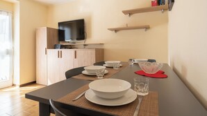 Un comedor con una mesa dispuesta para dos personas, un televisor sobre la mesa y una estantería con libros y una carpeta roja.