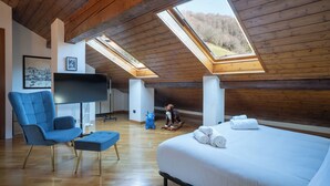 A bedroom with a large bed, a blue armchair, a TV, and wooden beams.