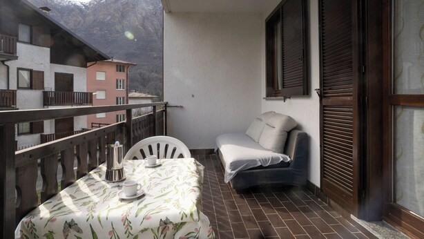 A balcony with a table set for two, a sofa, and a view of mountains. - ValleyHideaway with private garden near Como lake (Taceno)