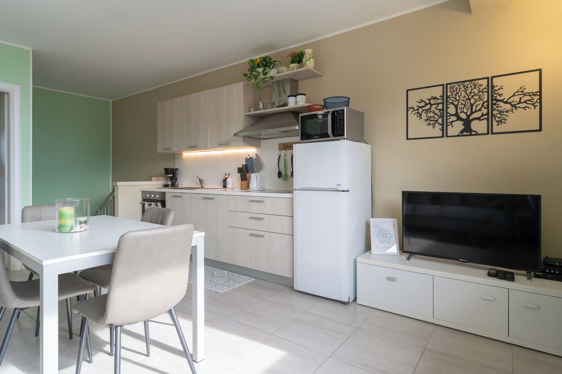A modern kitchen with white cabinets, a microwave, and a refrigerator. There is a dining table with chairs and a TV on a stand.