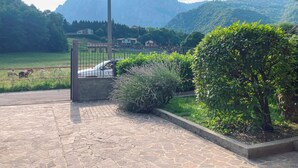 Une terrasse dallée de pierres, offrant une vue sur une chaîne de montagnes, et un espace clos où se trouvent une voiture et des vaches.