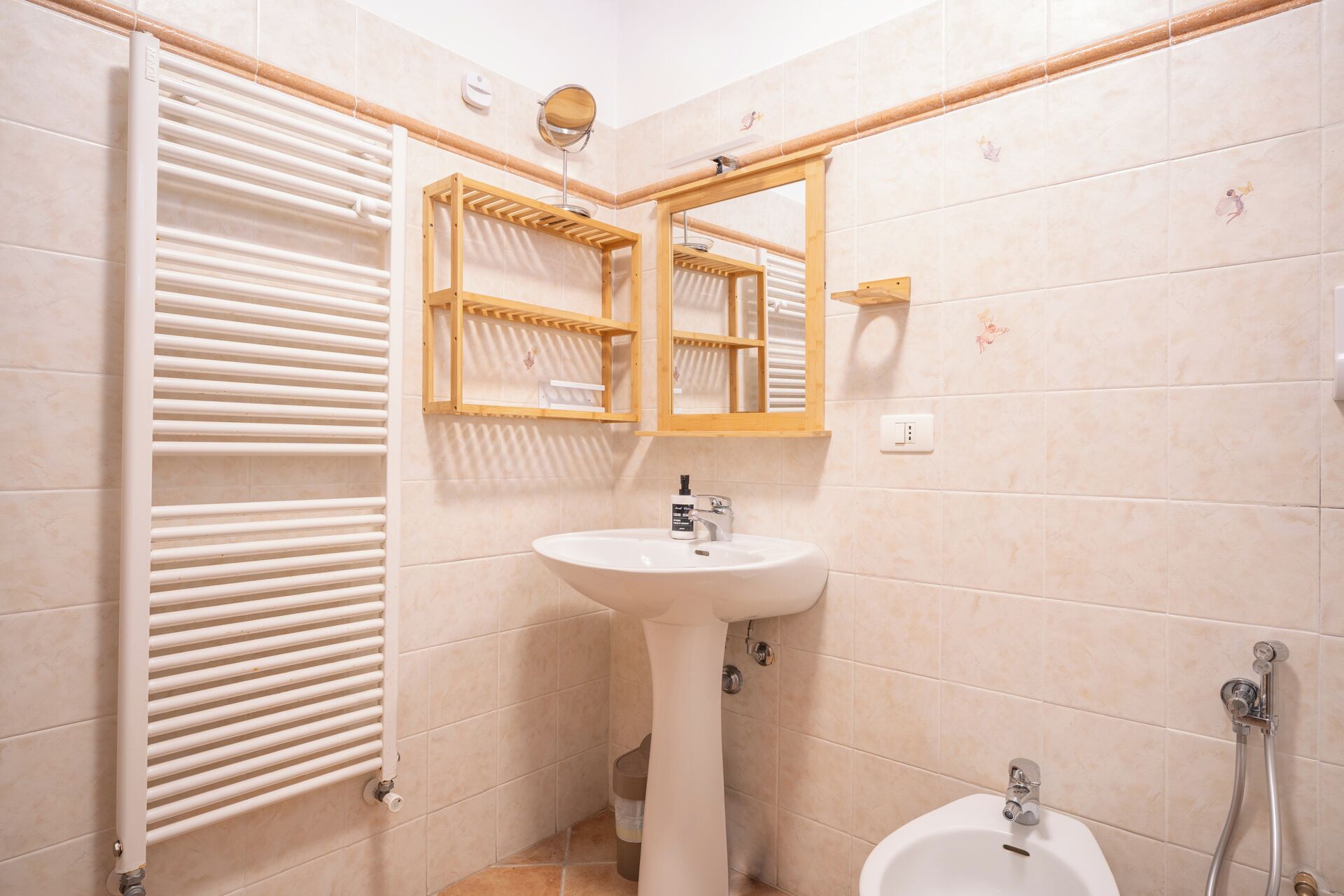 Un baño con un radiador blanco, un lavamanos pedestal, un estante de madera con un espejo y una zona de ducha.