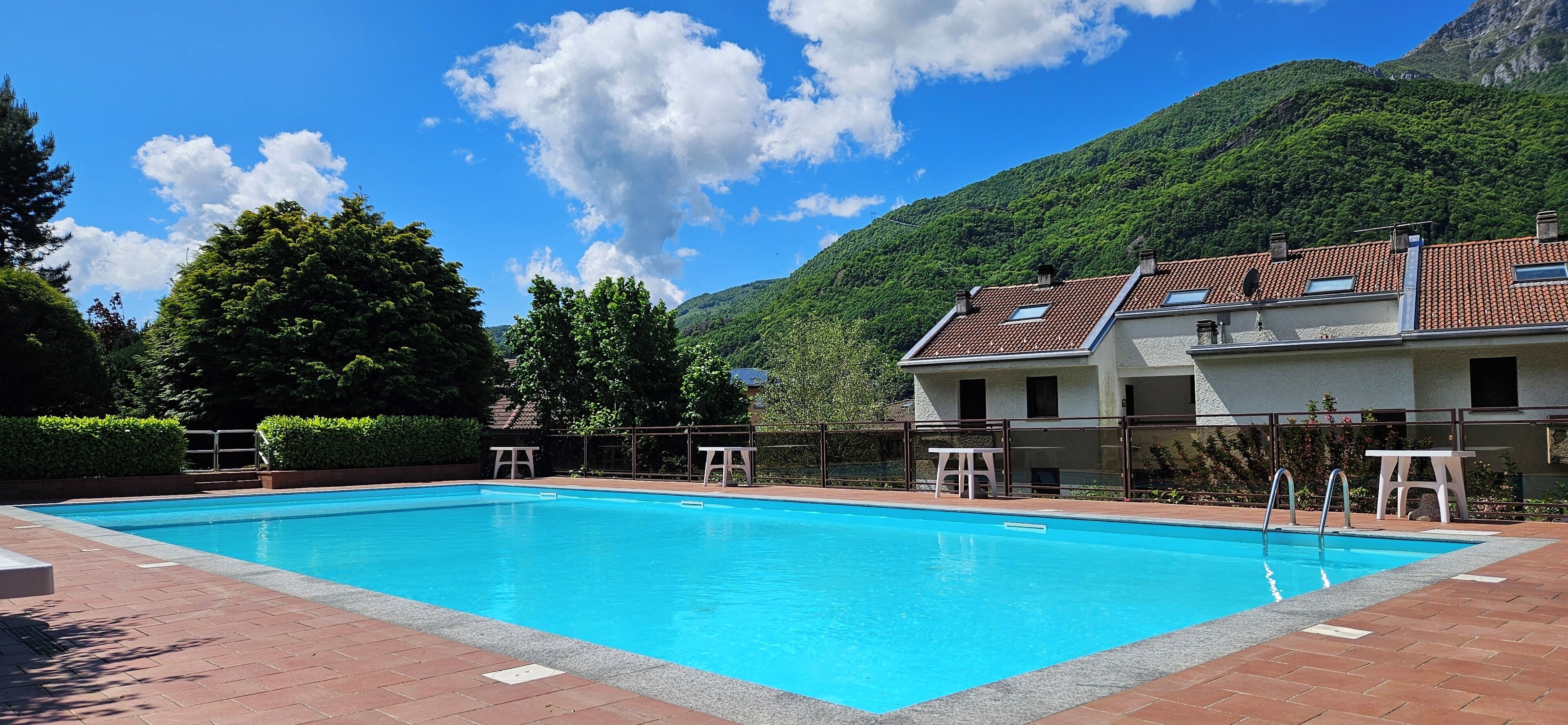 A rectangular swimming pool with a clear blue water surface, surrounded by a paved deck. There are two white sun loungers and a small white table near the pool. The pool is adjacent to a building with a red-tiled roof and a balcony.