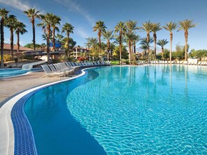 A large, curved swimming pool with clear blue water, surrounded by palm trees and lounge chairs.