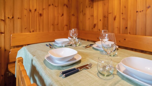 Une table à manger dressée avec de l’assiette blanche, des bols et des verres à vin, accompagnée d’un banc en bois et d’une nappe verte.